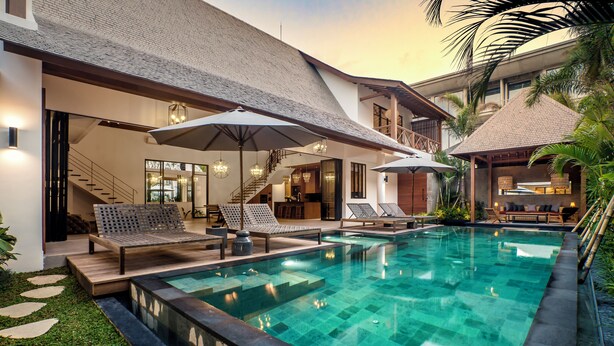 Un espace piscine moderne, doté de chaises longues et de parasols, entouré d’un jardin bien entretenu et d’un bâtiment à plusieurs niveaux avec un escalier.