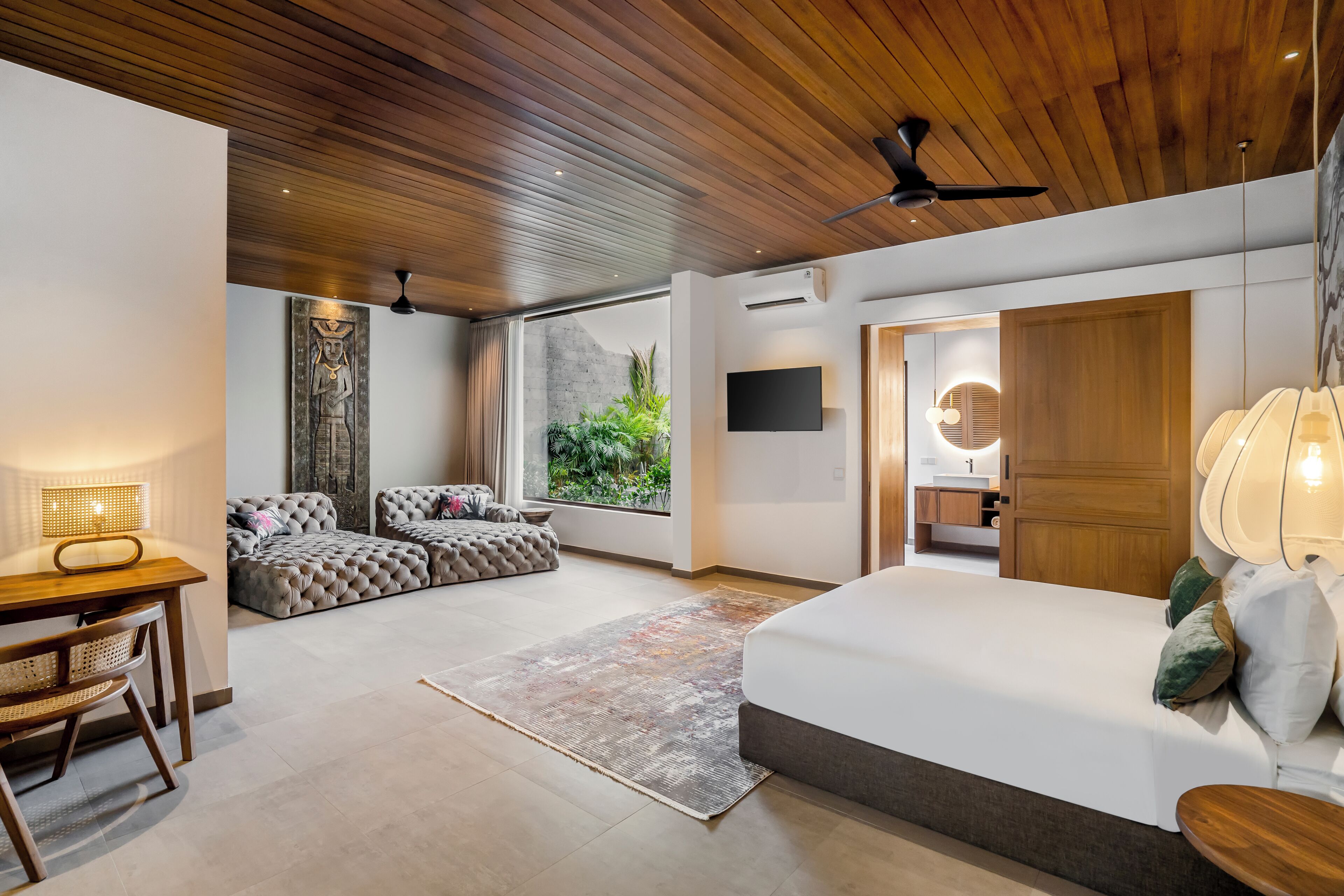 A modern bedroom with a wooden ceiling, a large bed, a seating area with a sofa and ottoman, a dining table with chairs, and a view of greenery through a large window.