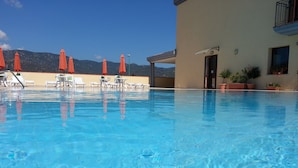 A clear blue pool with lounge chairs and orange umbrellas, surrounded by a building with a view of mountains. - Stylish Residence Le Fontane one bedroom apartment sleeps 4 persons no2503 (Villasimius)