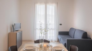 A modern living room with a dining area, a grey sofa, a TV on a cabinet, and a large window with sheer curtains. - Stylish Residence Le Fontane one bedroom apartment sleeps 4 persons no2503 (Villasimius)