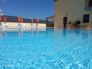 Piscina azul límpida com espreguiçadeiras e guarda-sóis laranja, rodeada por um edifício com vista para montanhas.