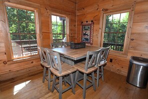 L’intérieur d’une cabane en bois, avec une table ronde et quatre chaises, de grandes fenêtres et une courtepointe accrochée au mur.