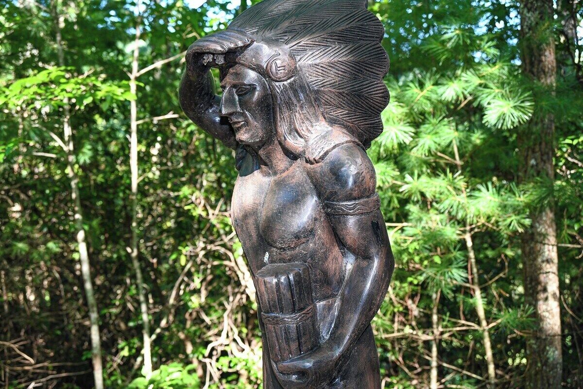 Une statue en bronze représentant une figure coiffée, placée sur un fond de feuillage.