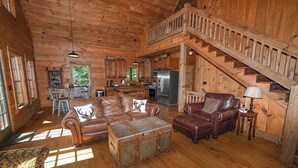 A cabin interior with a wooden staircase, leather furniture, and a kitchen area. - Mountain Lodge on 57 Acres • Hiking Trails • Private Lake • Hot Tub (Murphy)