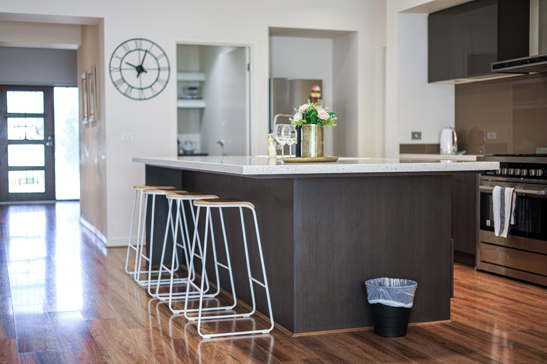 A modern kitchen with a central island, bar stools, a clock, and a trash bin.