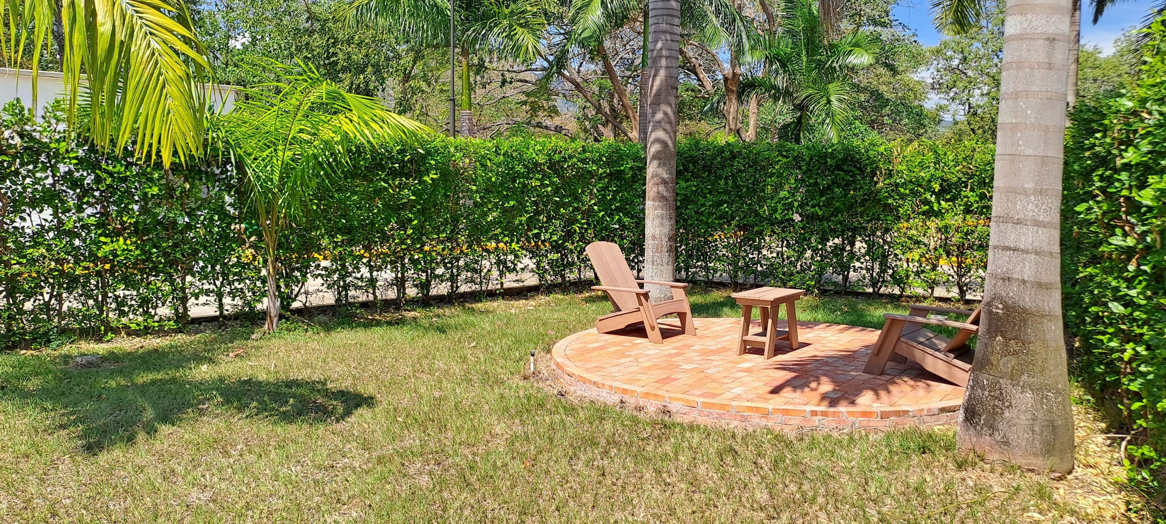 A circular patio with two wooden chairs and a small table, surrounded by lush greenery and palm trees.