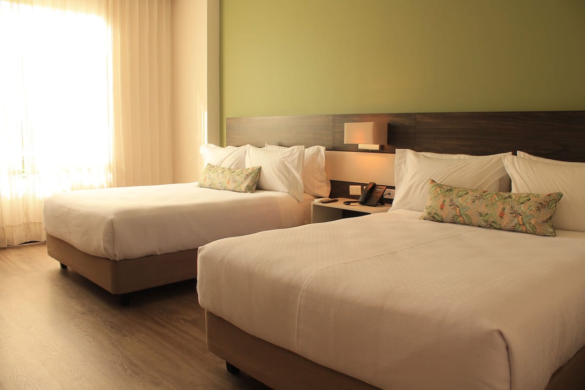 Two queen beds in a hotel room with a wooden floor, a nightstand, and a lamp.