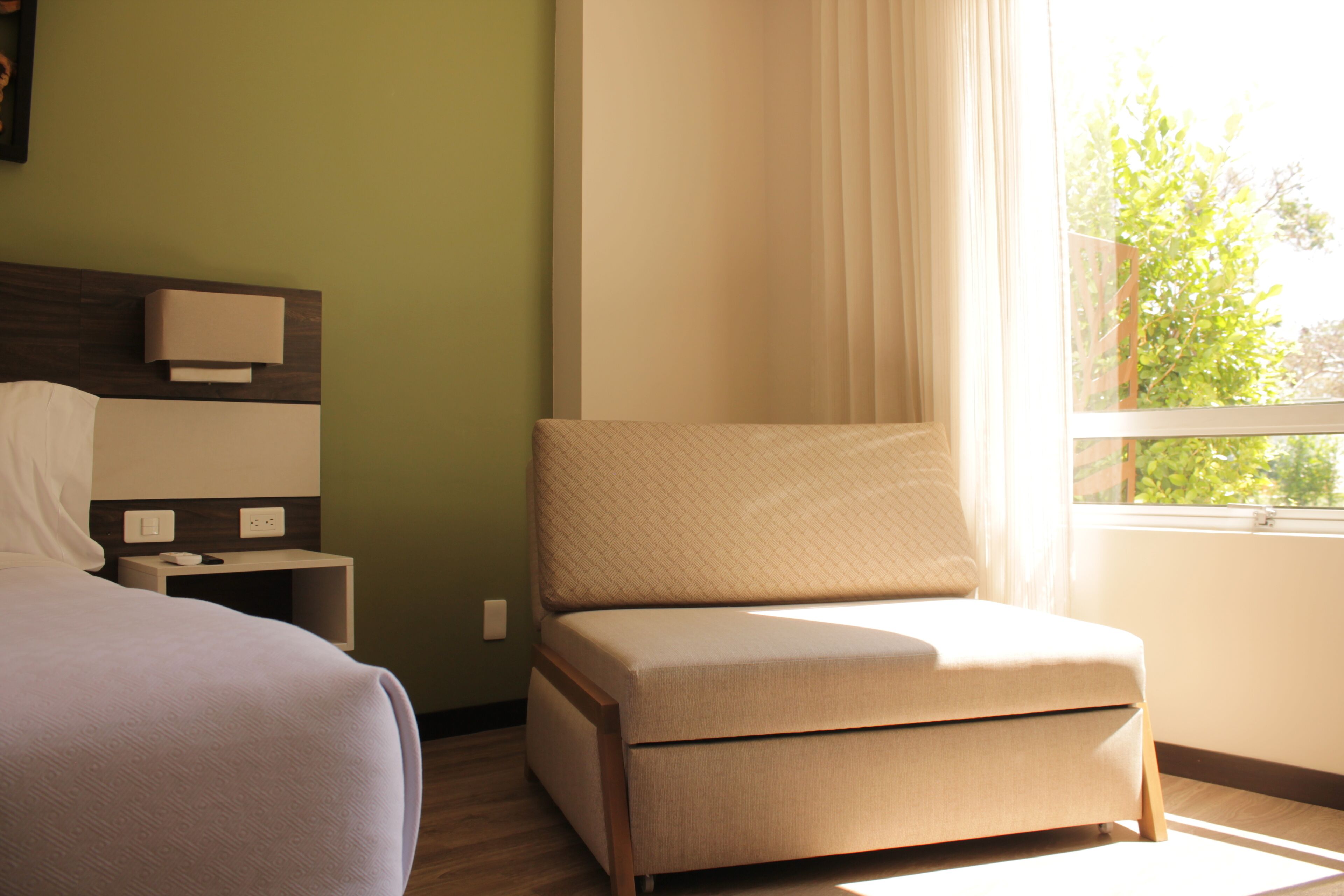 Habitación de hotel con cama, una mesita de noche con lámpara, una silla y un ventanal con cortinas.