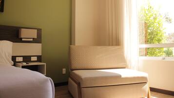 Habitación de hotel con cama, una mesita de noche con lámpara, una silla y un ventanal con cortinas.