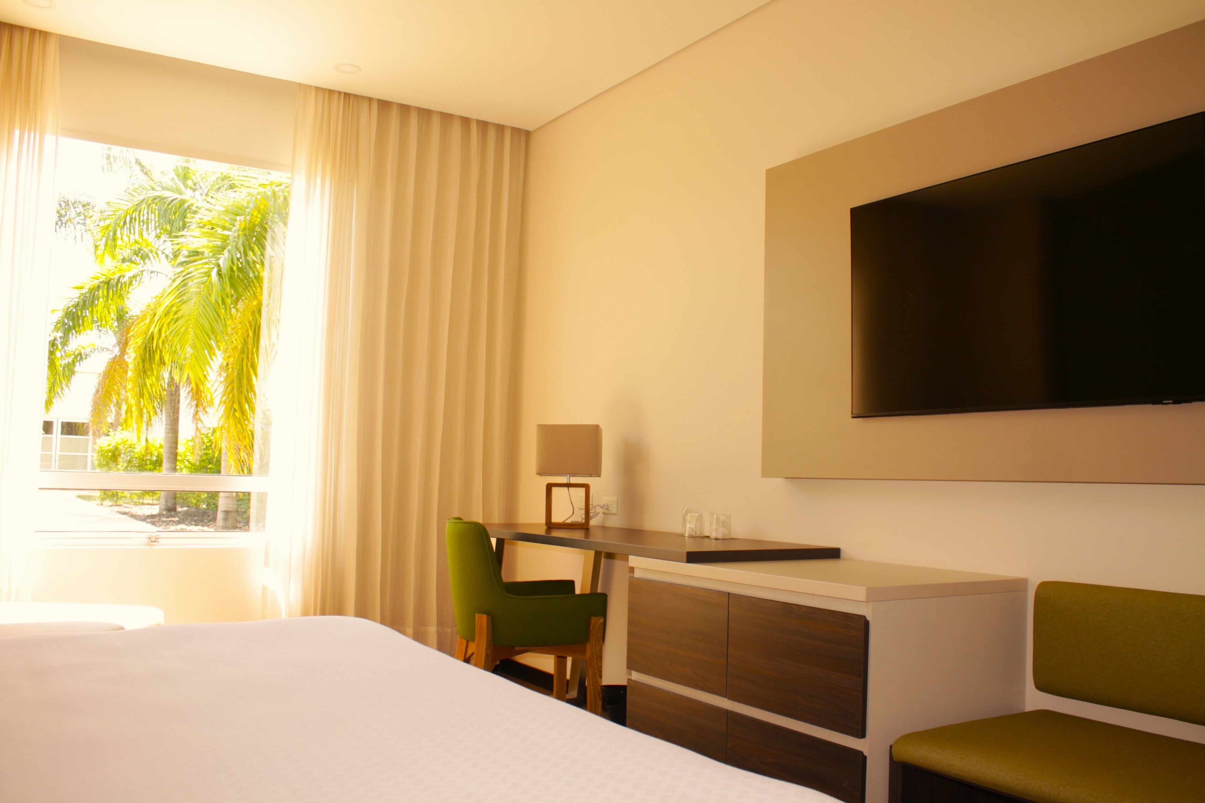 A hotel room with a bed, a desk with a lamp, a television, and a view of palm trees.