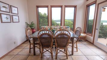 A dining area with a glass table and wicker chairs, large windows with a view, and framed pictures on the wall.