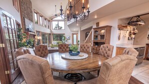 A spacious dining area with a large round table surrounded by six tufted chairs, a chandelier, and a view of a garden through large windows. - Lost Bridge Lodge on BEAVER LAKE! Across from POOL (Garfield)