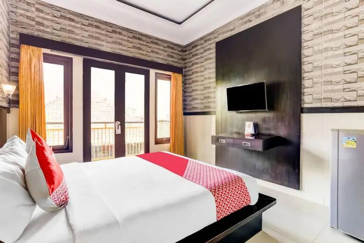 A hotel room with a bed, a flat-screen TV mounted on the wall, a minibar, and a balcony with curtains.