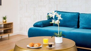 A modern living room with a blue sofa, a round coffee table with croissants and a glass of orange juice, and a potted orchid. - Merveil - Family Suite - Louvre - Sainte Anne (Paris)