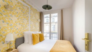A bedroom with a bed, a nightstand, a lamp, and a window with a view of buildings. - Merveil - Family Suite - Louvre - Sainte Anne (Paris)