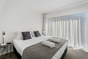 Una habitación de hotel moderna con una cama grande, una mesita de noche con lámpara y cortinas translúcidas.