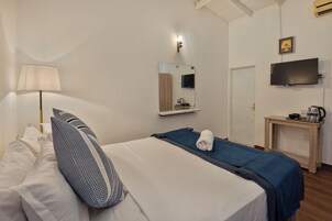 Habitación de hotel con cama, mesita de noche con lámpara, espejo, televisión y una pequeña mesita de madera con tetera y tazas.