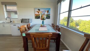 A dining area with a glass-top table, wooden chairs, a vase with flowers, a painting of a sunset, and a view of trees through a large window. - Morwell Pet friendly 3BM House (Morwell)