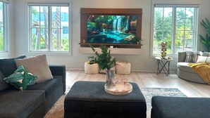 Una sala de estar moderna con un televisor de pantalla plana grande montado en la pared, un sofá gris, una mesa de centro con una bandeja de vasos y ventanas amplias que ofrecen vistas a la vegetación exterior.