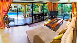 A spacious living area with a large sofa, a wooden coffee table, and a TV. There is a dining table with chairs and a view of a swimming pool through large glass doors. - Tropical pool villa, perfect for big families, group getaways & family reunions (Tambon Rawai)