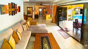 A spacious living area with a sofa, coffee table, and decorative rugs. A dining area with chairs and a table is visible through a glass door. - Tropical pool villa, perfect for big families, group getaways & family reunions (Tambon Rawai)