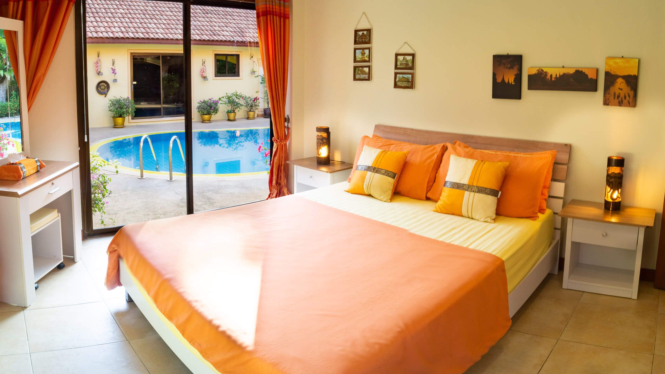 A bedroom with a large bed, a nightstand, a bedside table, a window with curtains, a wall with framed pictures, and a view of a swimming pool outside.