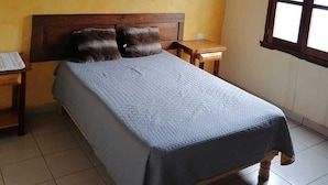 Un dormitorio con una cama, cabecera de madera, dos mesitas de noche, un ventilador de techo y una ventana con persianas.