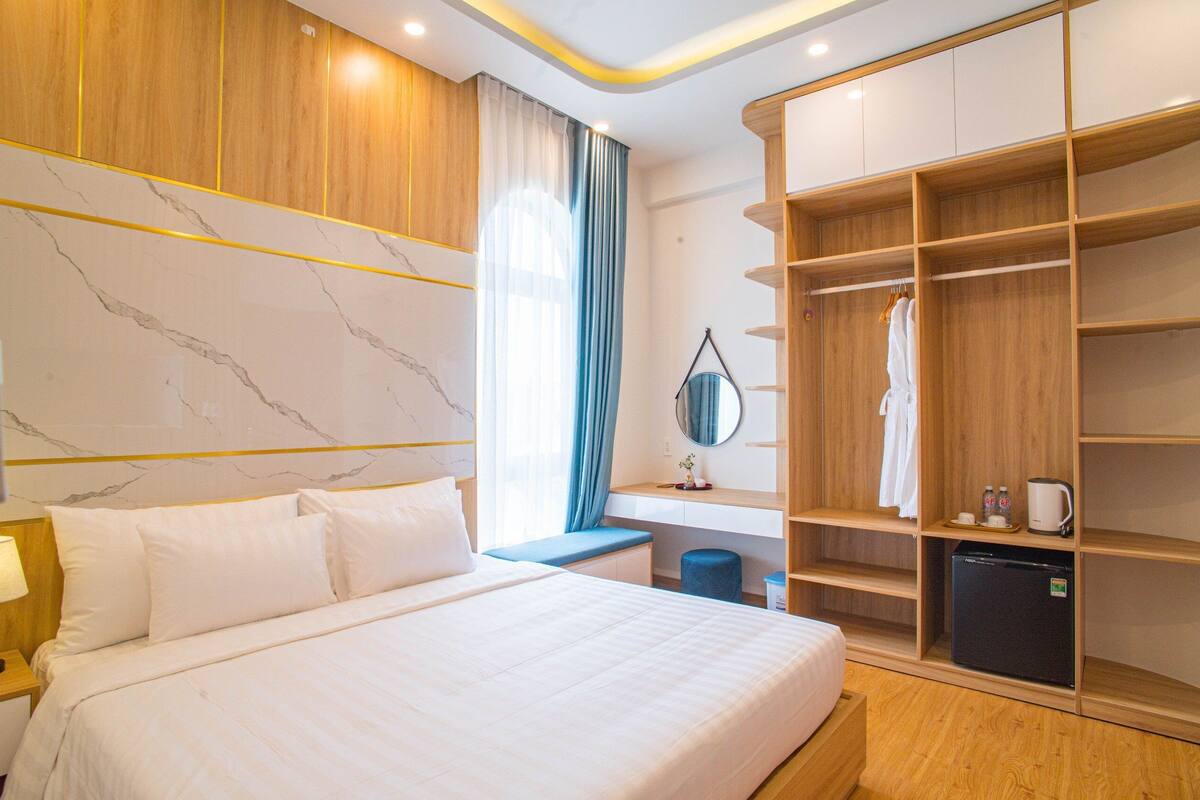A hotel room with a large bed, a wooden wardrobe, a desk with a mirror, and a small refrigerator.