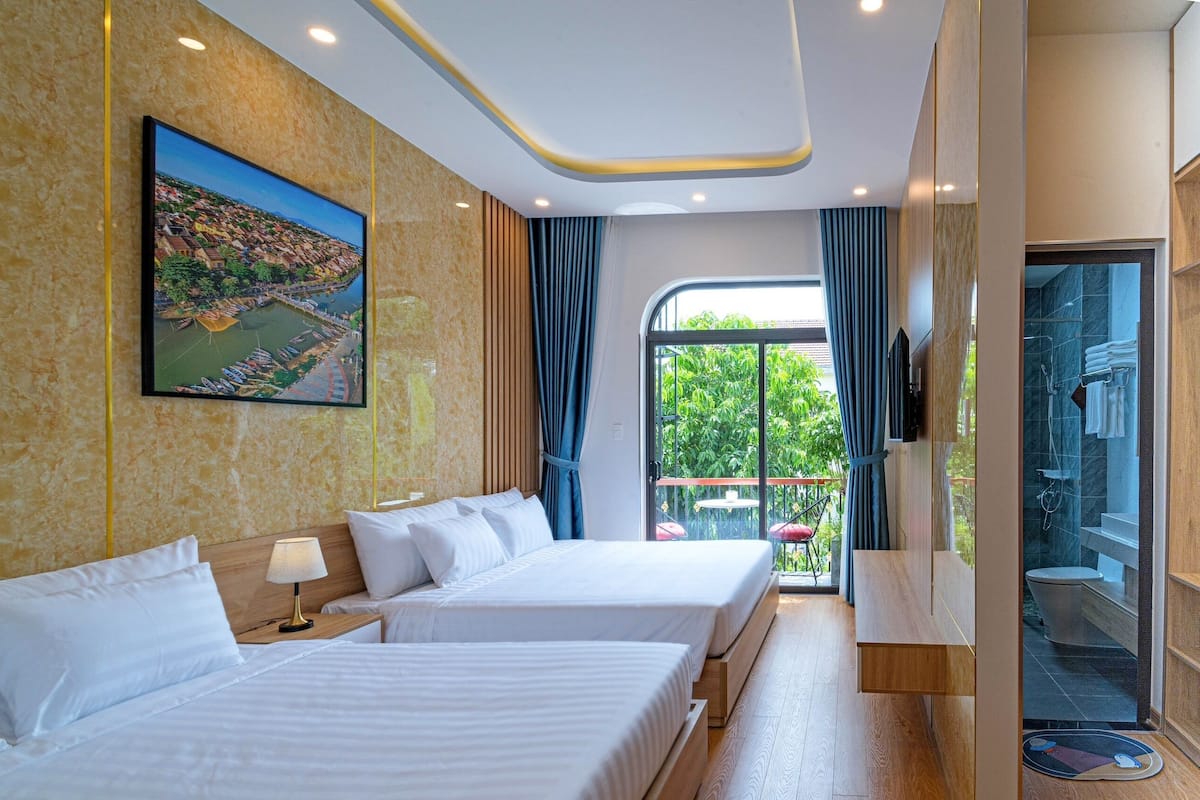A hotel room with two beds, a large window with a view of greenery, and a bathroom with a glass door.