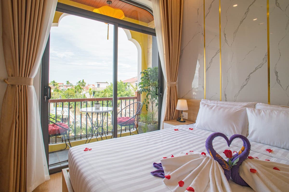 A hotel room with a large balcony, a bed with a heart-shaped towel, and a view of palm trees and buildings.