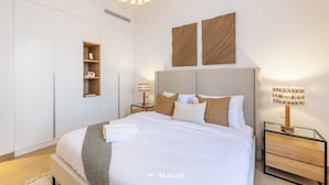 A modern bedroom with a large bed, built-in wardrobe, bedside tables, and decorative wall art. - Manzil - 1BR | Near La Mer Beach | Pool & Sea View (Dubai)