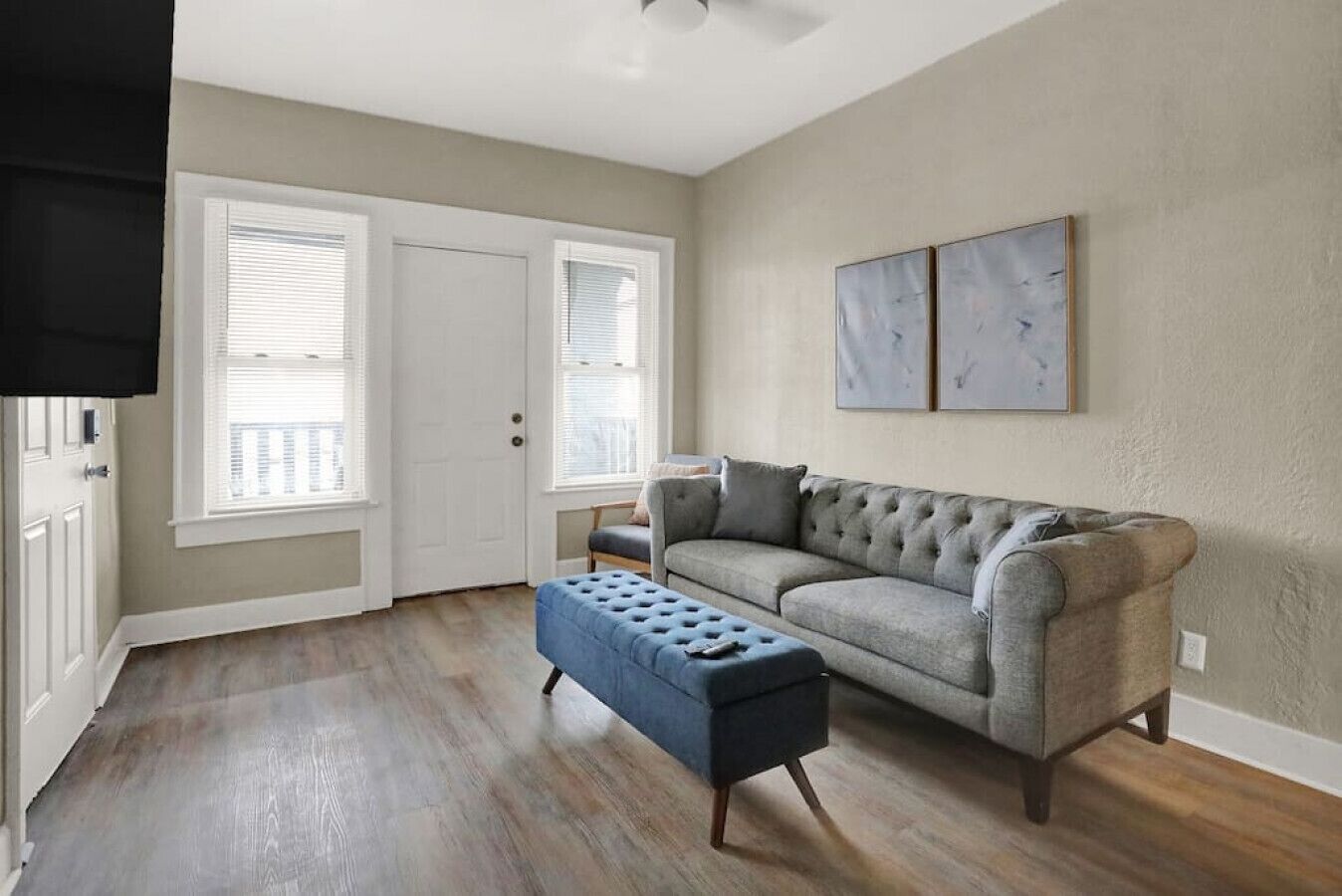 A living room with a grey sofa, a blue ottoman, and a door with white blinds.