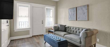 A living room with a grey sofa, a blue ottoman, and a door with white blinds.