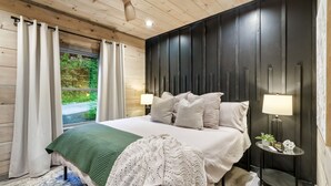 A bedroom with a bed, bedside lamps, a small table with a lamp, and a view of greenery outside.