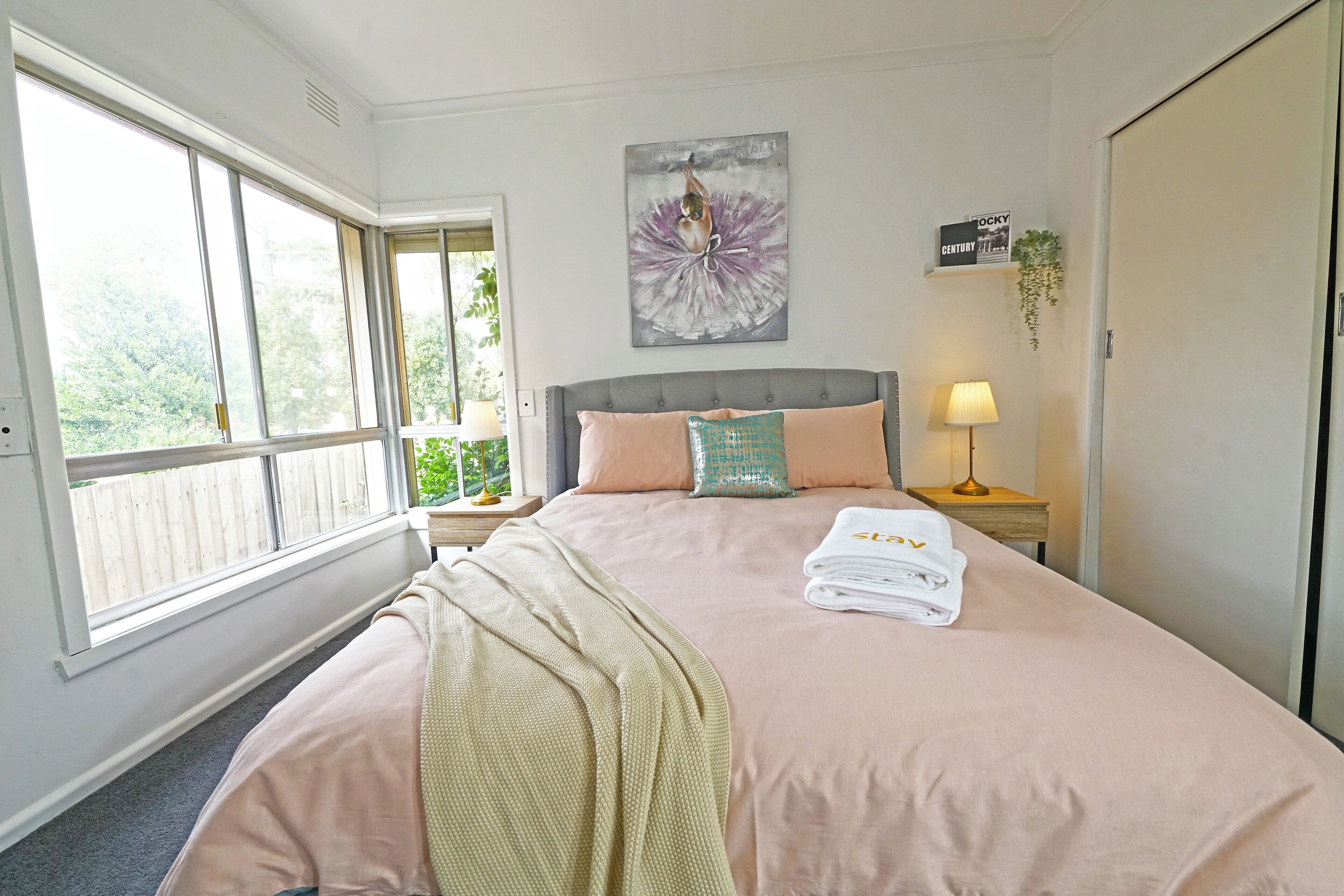 Un dormitorio con un ventanal grande, una cama con colcha rosa, cabecero gris y un cuadro en la pared.
