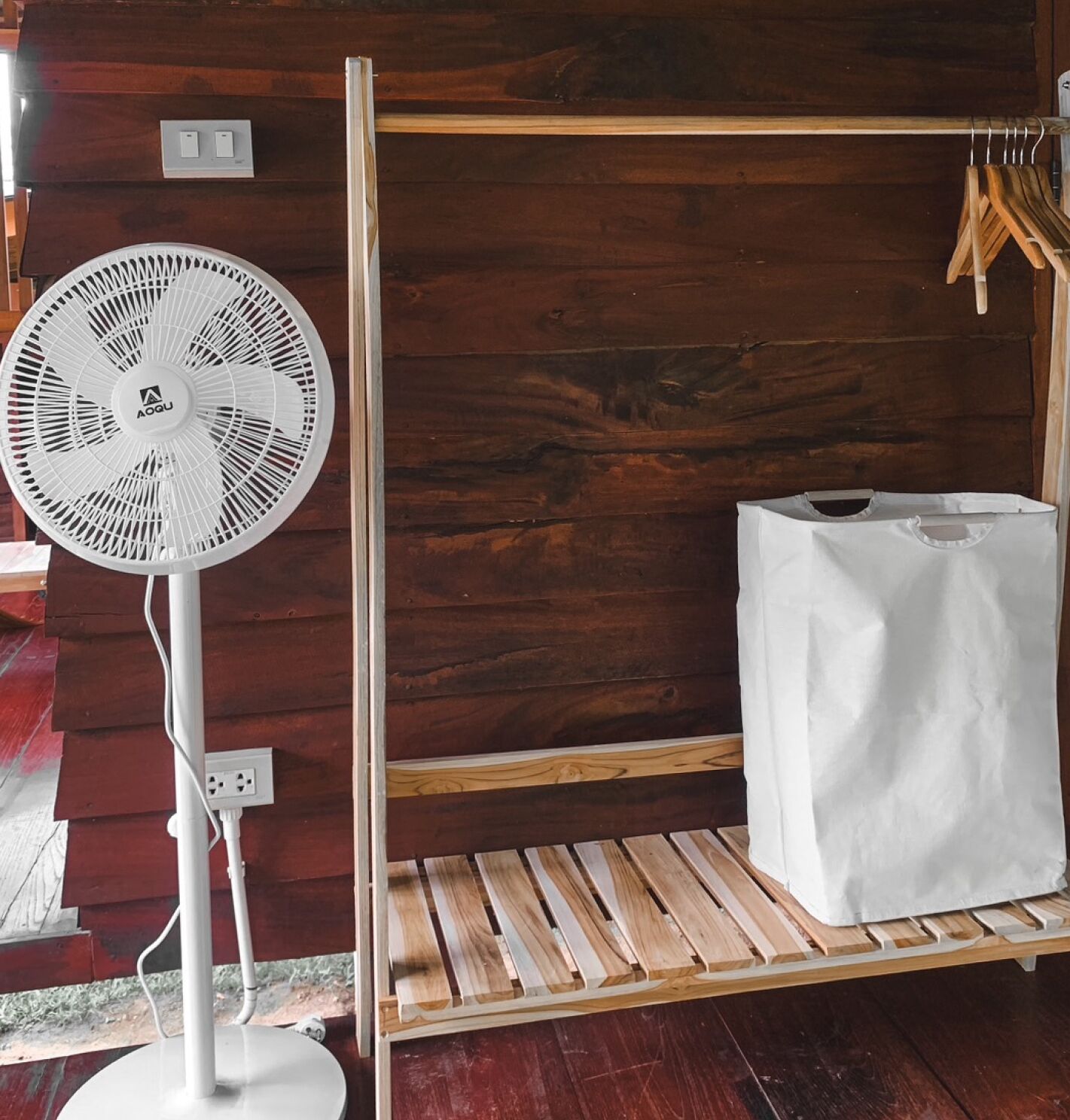 Una habitación con un ventilador de pie blanco, un estante de madera y una bolsa blanca sobre un banco de madera.