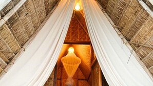 Una habitación acogedora y rústica con techo de paja, una cama con cojines verdes, una lámpara colgante y una botella de vino en el suelo.