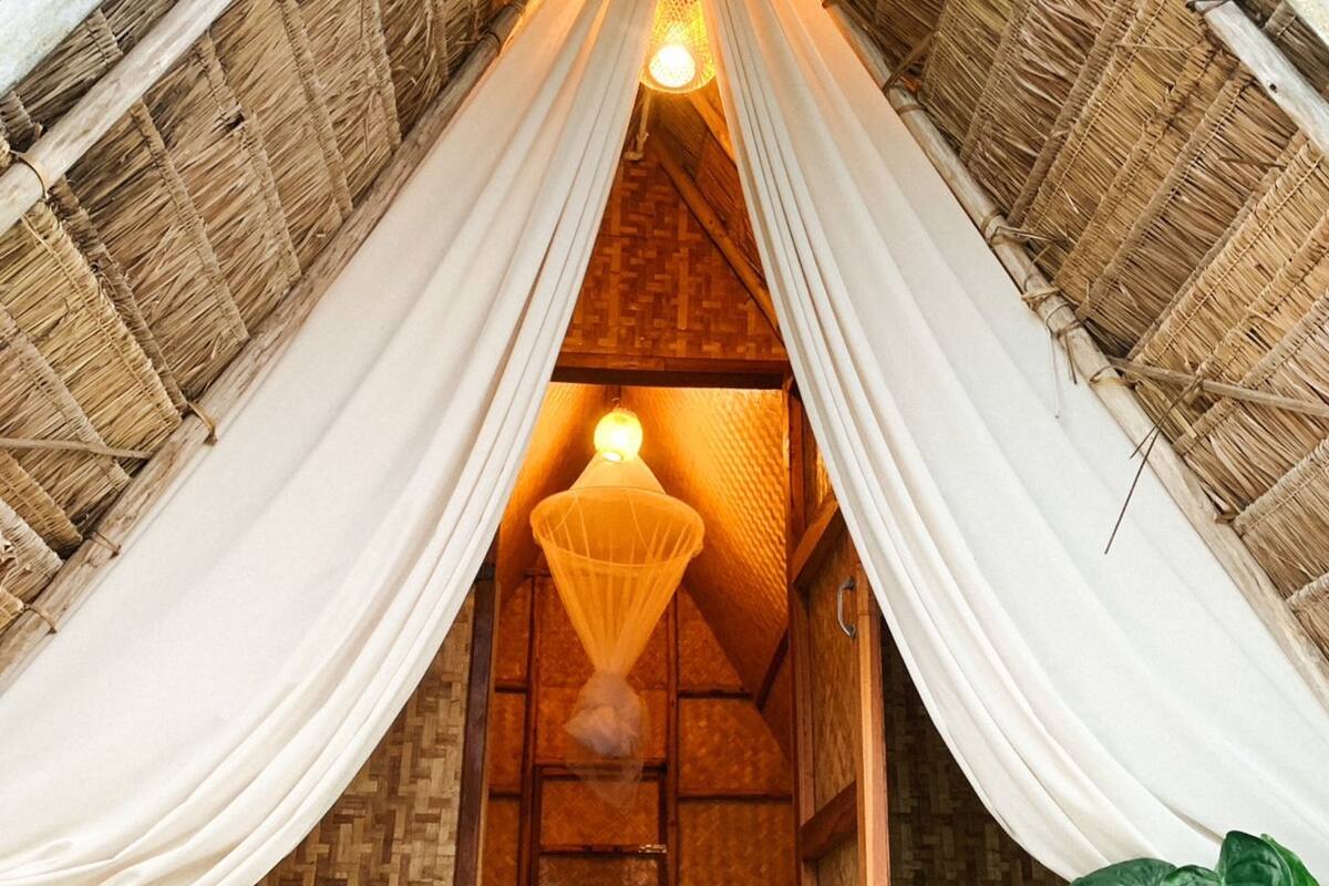 Une pièce chaleureuse et rustique, avec un toit de chaume, un lit agrémenté de coussins verts, un luminaire suspendu et une bouteille de vin posée sur le sol.