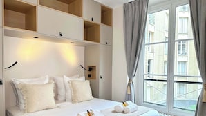 A hotel room with a bed, built-in shelves, a window with curtains, and a view of buildings outside.