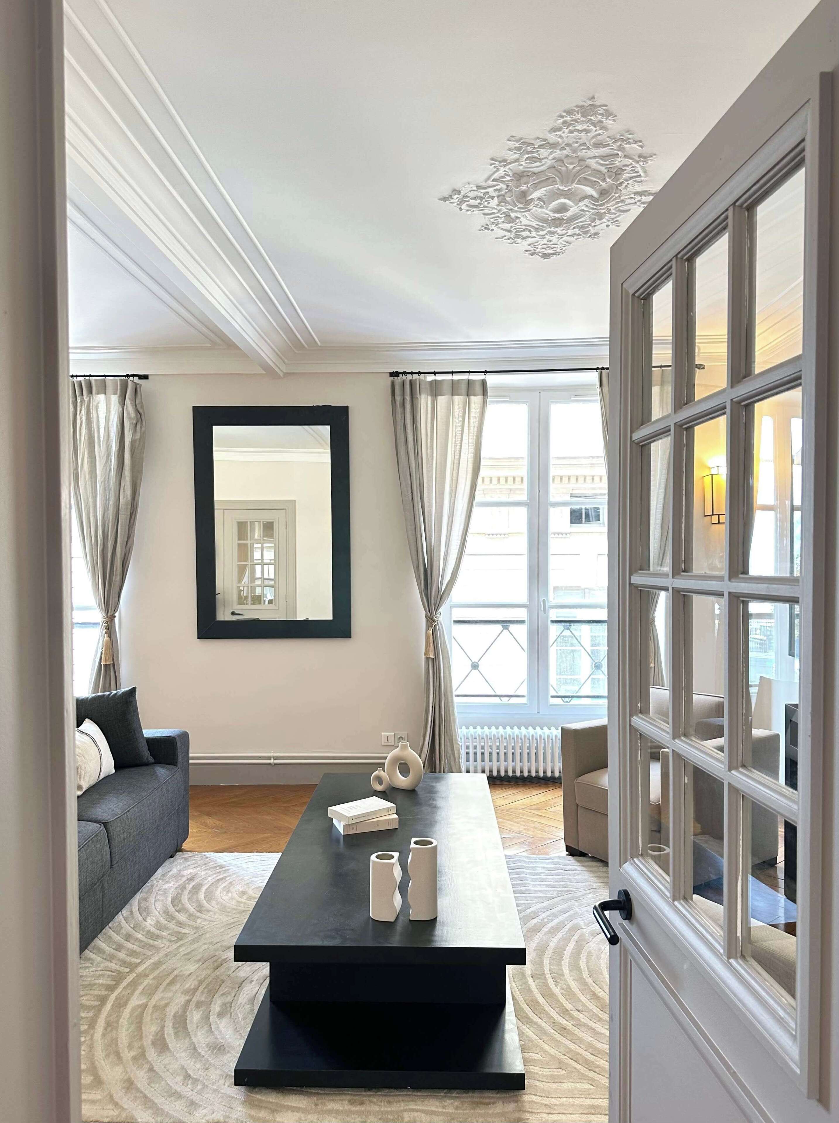 A modern living room with a black coffee table, a sofa, a large mirror, and a view of the city through large windows.