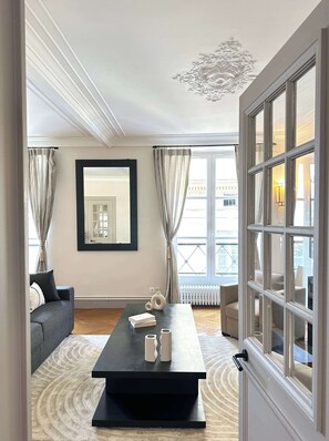 A modern living room with a black coffee table, a sofa, a large mirror, and a view of the city through large windows.