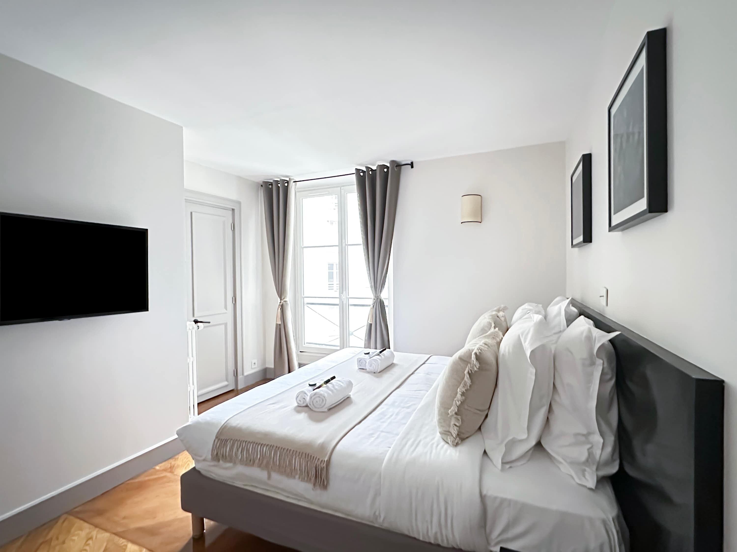 A modern bedroom with a bed, a flat-screen TV, a window with curtains, and a wall-mounted light fixture.