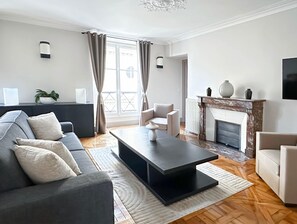A modern living room with a sofa, a coffee table, a fireplace, and a view through a window. - Luxembourg Gardens Prestige flat for 8 lift  4 BR (Paris)