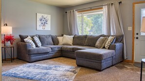 Ein Wohnzimmer mit grauem Ecksofa, einem blau-beigen Teppich, einer gerahmten Blumenarbeit und einem Fenster mit Vorhängen.