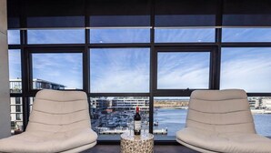 Ein Zimmer mit zwei beigefarbenen Sesseln, die auf ein großes Fenster mit Blick auf einen Yachthafen und Gebäude zeigen.
