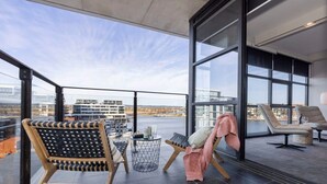 Ein moderner Balkon mit Blick auf eine Stadtlandschaft, ausgestattet mit einem Holzstuhl, einem kleinen runden Tisch und einer Liege mit Decke.