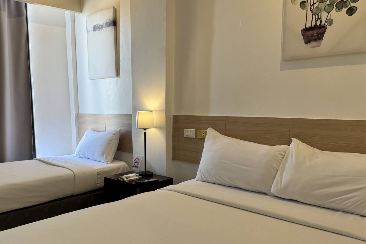 Una habitación de hotel con dos camas individuales, una mesita de noche con lámpara, un cuadro en la pared y una ventana con cortinas.