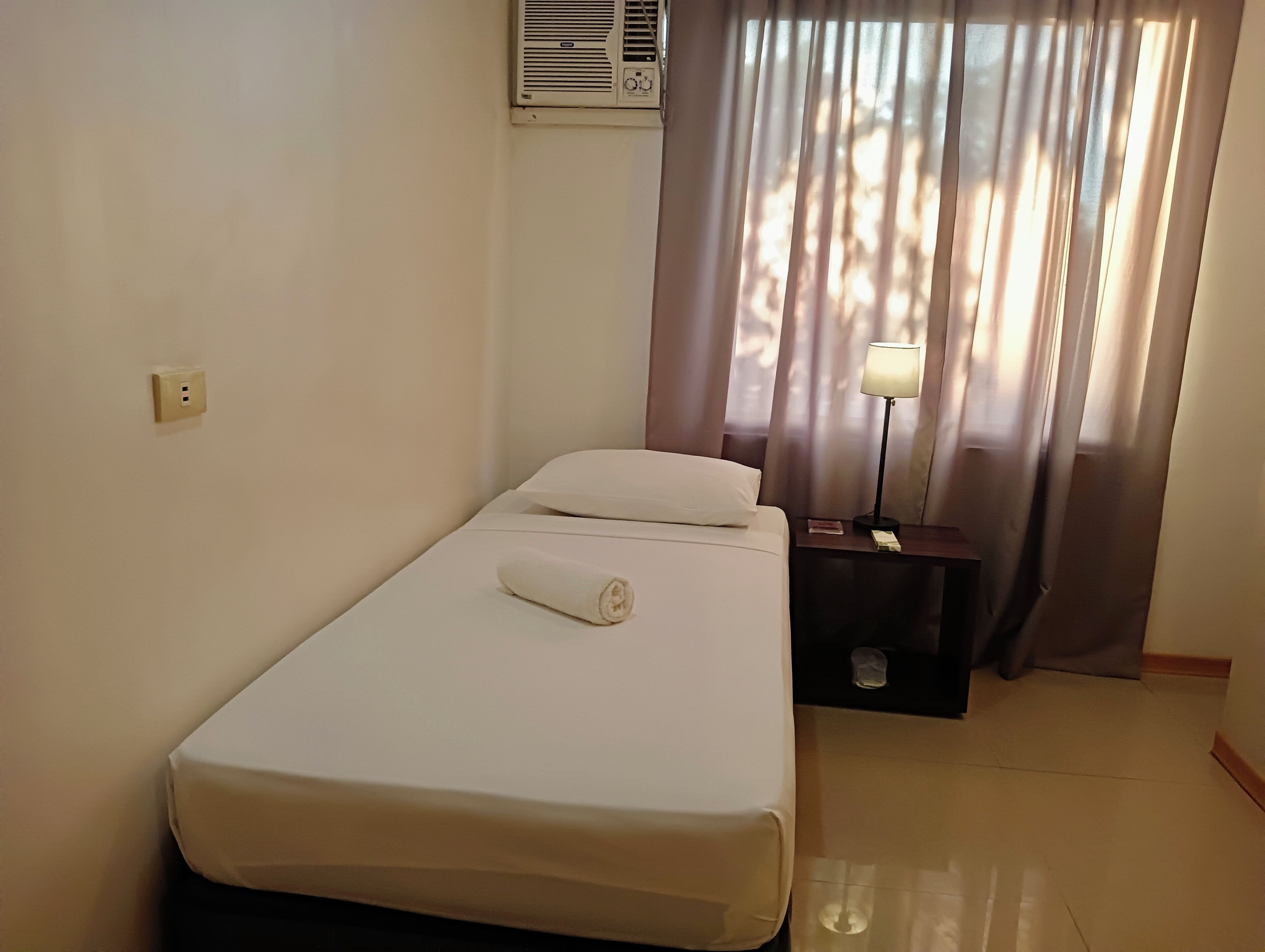 Uma cama de solteiro com colchão e travesseiro brancos, uma mesa de cabeceira com abajur e uma janela com cortinas.