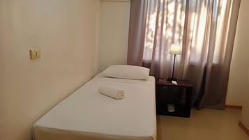 Uma cama de solteiro com colchão e travesseiro brancos, uma mesa de cabeceira com abajur e uma janela com cortinas.