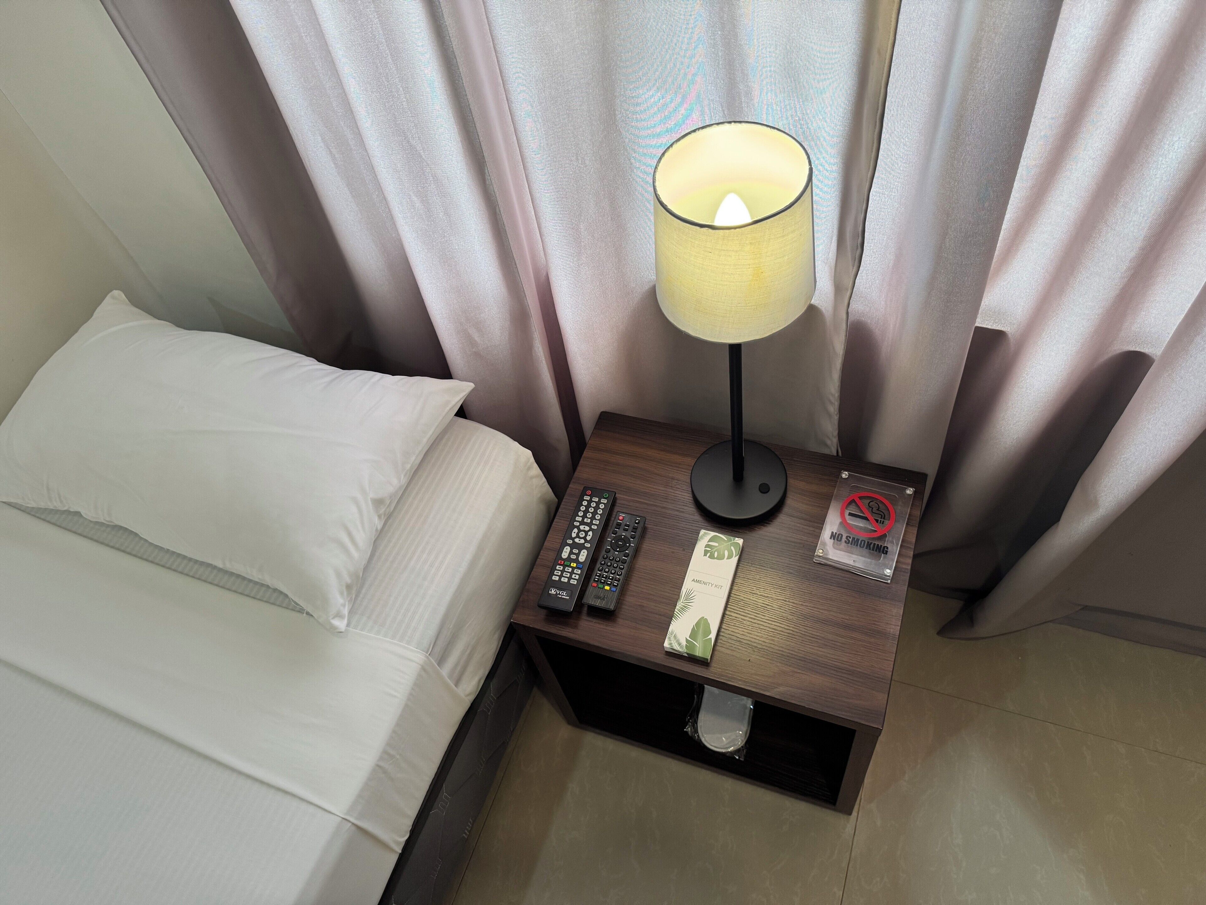 Habitación de hotel con cama, mesita de noche con lámpara, control remoto, cartel de no fumar y set de amenidades.